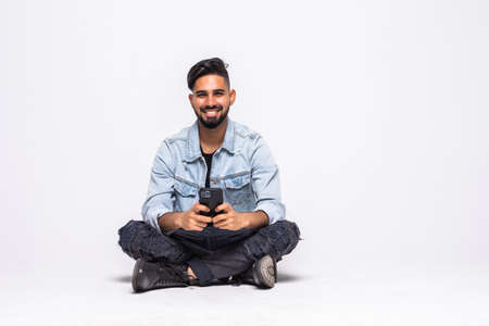 Cheerful young man sitting with legs crossed isolated over white background, using mobile phone