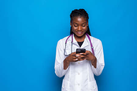 Young african woman doctor posing isolated over blue wall background using mobile phone.の写真素材