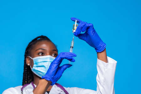 Syringe and tube in hand of african american doctor in medical mask on blurred background isolated on blue backgroundの写真素材