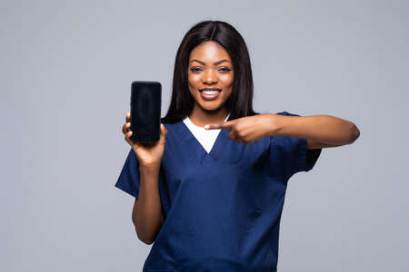 African american doctor woman isolated on white background. Female doctor in medical gown pointing on mobile phone with blank empty screen. Healthcare personnel medicine concept.の写真素材