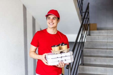 Image of smiling young delivery man in red uniform holding pizza boxes and coffee takeaway at city...