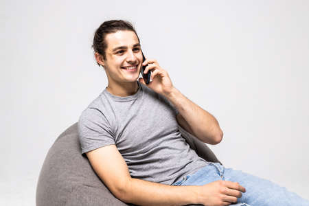 Handsome young man wearing a casual outfit, sitting on a bean bag, holding his phone talking to someone, looking away, isolated on white backgroundの写真素材