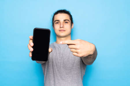 Young man pointing on smartphone with blank screen, showing application against blue backgroundの写真素材