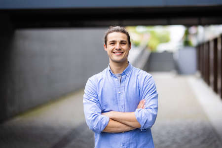 Handsome smiling young man with folded arms. Beautiful portrait of laughing joyful cheerful men with crossed hands in a cityの写真素材