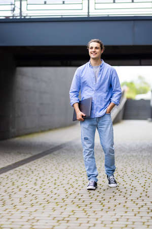 Portrait of a young serious man holding laptop computer while standing in a city area