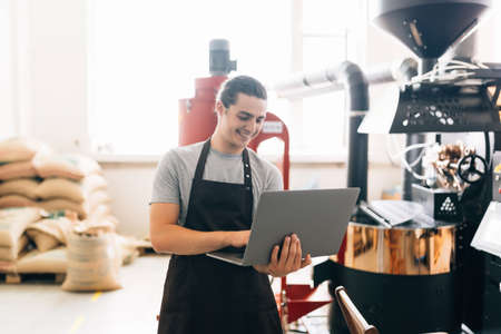 Young master working with special equipment. Man controlling brown beans pouring into cooler machine from large coffee roaster while using laptop for the manage the coffee machineの写真素材