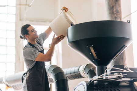 Professional coffee roaster worker wearing apron holding heavy bucket with coffee beans in hands while standing at the factory. Industry conceptの写真素材