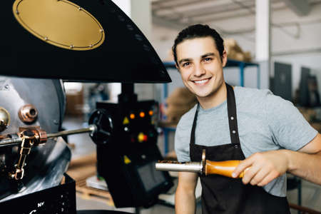 Handsome man in apron looking at freshly roasted coffee beans while standing by coffee roasting...