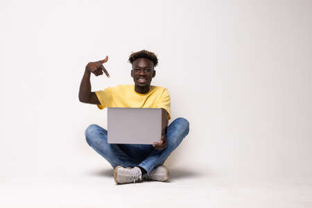 Young black man sitting on the floor with a laptop person pointing by hand to a shirt copy space, proud and confidentの写真素材