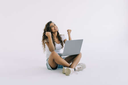 Woman sitting on the floor with a laptop computer and arms up on white background