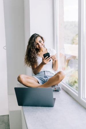 Young woman sitting on a windowsill with notebook and talking on mobile phone. Girl working at home,...