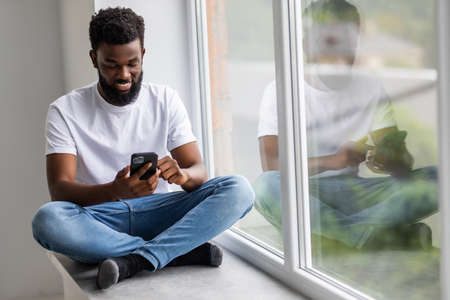 Handsome african man sits home on windowsill, reads text messages, sends email, uses his phoneの写真素材