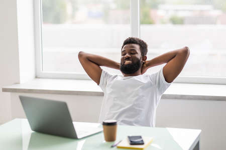 Cheerful african man watching at his laptop screen, at his work place, with arms behind the head, resting, smiling, in the officeの写真素材