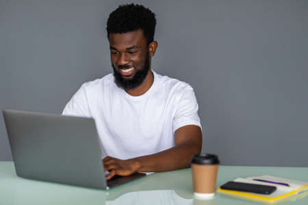 Young african-american male sitting at desk with laptop tidy office in smiling and looking at camera