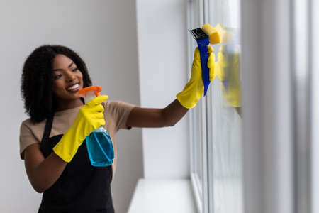 Beautiful african woman is using a duster and a spray, looking at camera and smiling while cleaning windows in the houseの写真素材