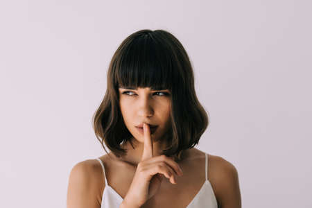Portrait of a confident woman showing silence gesture and looking at camera isolated over white...