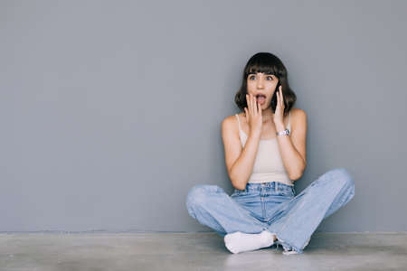 Young woman wearing eyeglasses sitting on floor while talking by phone over gray background. Look aside.の写真素材