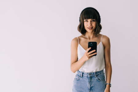 Young beautiful girl in headphones smiling looking at phone screen over white background.の写真素材