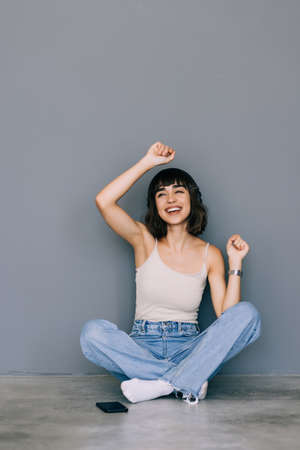 Pleased woman wearing casual clothing singing while listening to music via wireless earphones and sitting on floor with legs crossed on gray backgroundの写真素材