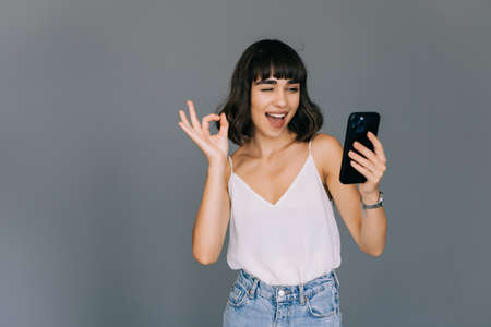 Attractive woman showing ok gesture while holding mobile phone over gray backgroundの写真素材