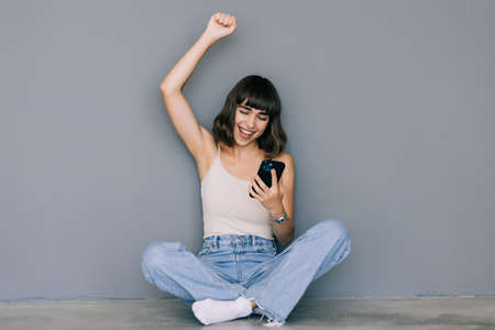 Portrait of a smiling woman win sitting on the floor and talking on the phone on gray backgroundの写真素材