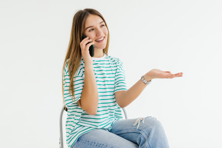 Happy young woman talking on smartphone, sitting in armchair against white studio wall, empty space.の写真素材