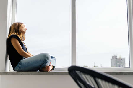 Beautiful young woman with cup of coffee sitting on windowsillの写真素材