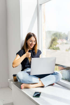 Online communication. Young woman sitting on windowsill at home and talking to family, friend or...