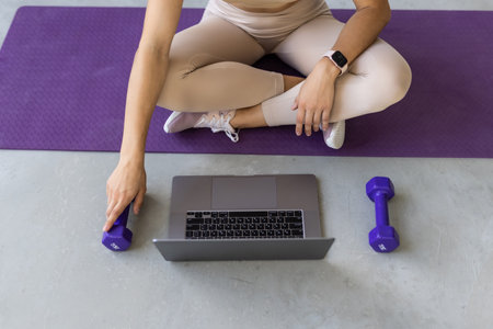 Sporty woman in sportswear is sitting on the floor with dumbbells is using a laptop at home in the living room. Sport and recreation concept.の写真素材