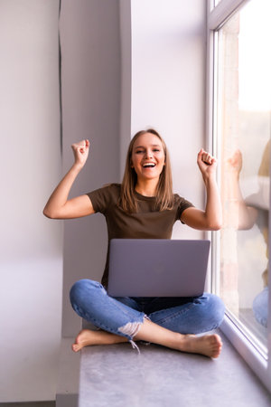Young pretty woman sitting near the window with a laptop with raised hands of good newsの写真素材
