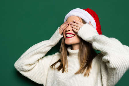 Young beautiful woman wearing Christmas Santa hat over isolated green background covering eyes with hands smiling cheerful and funny. Blind concept.の写真素材