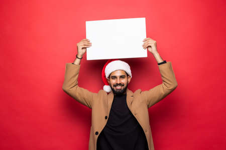 Young indian man in santa hat holding white blank board standing on red background. Christmas mood.の写真素材
