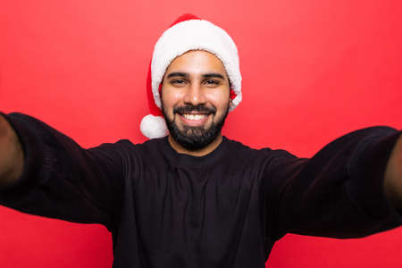 Handsome young man wearing santa hat, taking selfie standing over red backgroundの写真素材