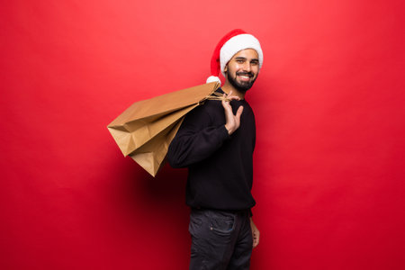 Young handsome man in santa hat holding shopping bag on red background