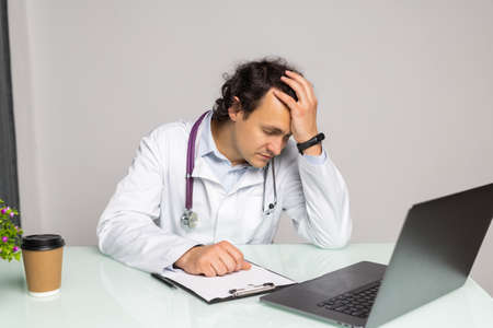 Portrait of tired doctor in glasses sitting at the desktop. Physician is working in bright office, medicine concept, copy spaceの写真素材