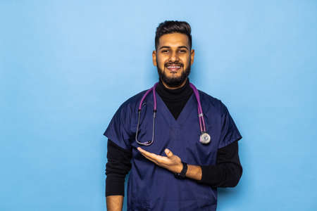 Smiling young male doctor showing and presenting something with hand, isolated on blue background...