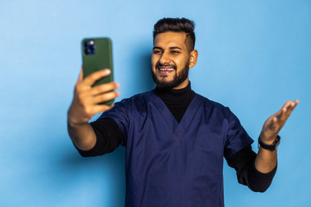 medicine, healthcare and technology concept - happy smiling indian doctor or male nurse in blue uniform with stethoscope having video call on smartphone over white backgroundの写真素材