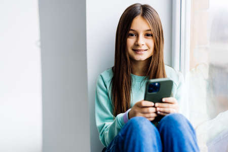 Pretty teenage girl sitting on windowsill with smartphoneの写真素材
