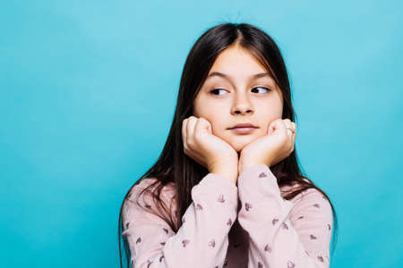 Dreamy beautiful caucasian little girl wearing blue T-shirt over blue background with pleasant expression, looks sideways, keeps hand under chin, thinks about something pleasant.の写真素材