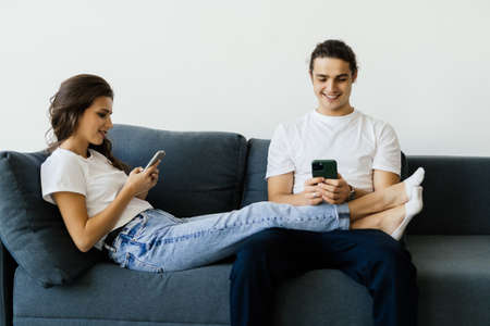 Mobile addiction concept, young couple ignoring each other using smartphones relaxing on sofa,...