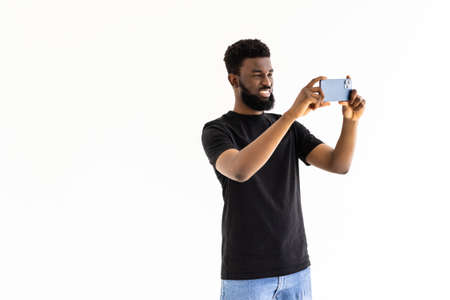 Portrait of young handsome African American man standing against gray background using smartphone to take selfie pictures and smilingの写真素材