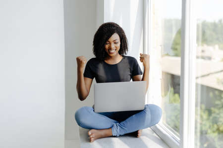 African Americanwoman with laptop celebrate win sitting on windowsill