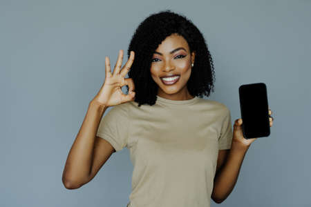 Portrait of smiling young african woman holding blank screen mobile phone and showing ok over gray...