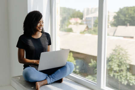 Beautiful young african woman sitting on the windowsill with a laptop.の写真素材