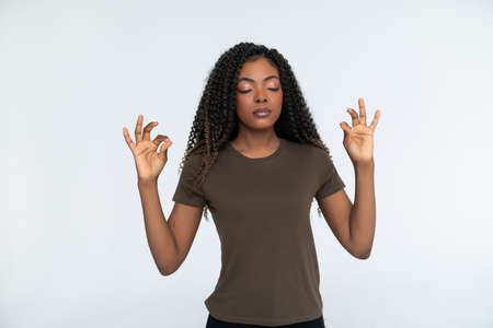 consideration and prayer. Beautiful calm young black female keeping eyes closed while practicing yoga indoors, meditating, holding hands in mudra gesture, thinking about peaceの写真素材