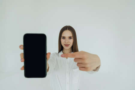 Portrait of an excited young business woman pointing finger at blank screen mobile phone isolated over white backgroundの写真素材