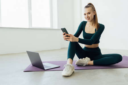 Young woman in sportswear sitting floor resting while doing fitness yoga at home using phone chatting.の写真素材