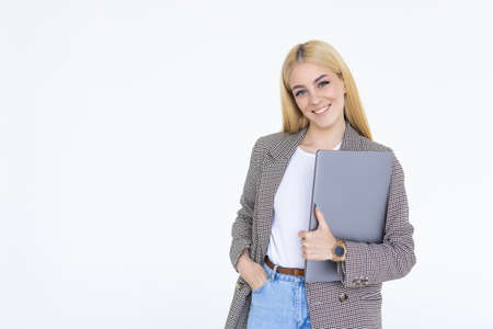 Happy woman in clothes looking at camera with smile and browsing laptop while working against white...
