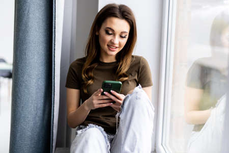 Attractive young woman sitting on a windowsill at home and texting on her phone