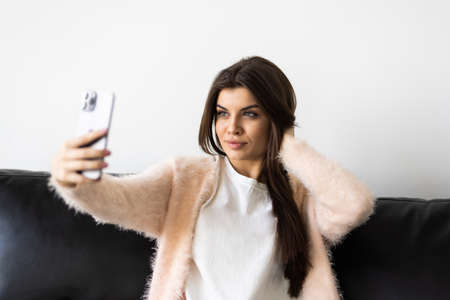 Photo of brunette woman 30s taking selfie on cell phone while sitting on couch in bright apartmentの写真素材